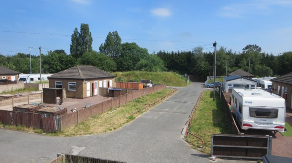 A photo of a small brick building and a white caravan with a tarmac drive and grass separating them 