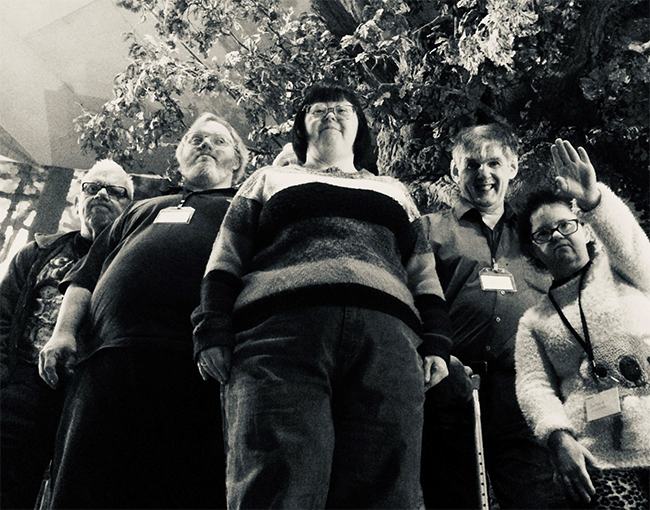A group of five men and women artists from The Professors stand in front of a big replica tree at Weston Park Museum. They look down into the camera. 