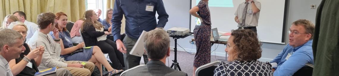 Photo of people in a room sitting down and chatting with a picture of a map on the wall in front