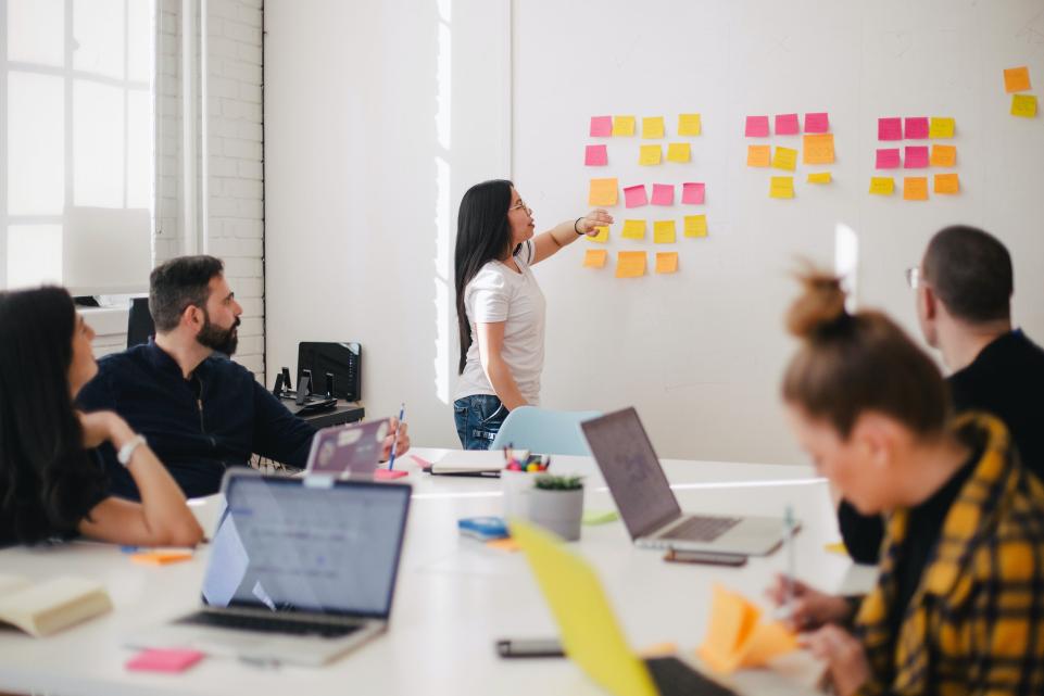 Group of people sat around a white desk, they have laptops out and also have post-it notes on a white board at the front of the room where one woman is stood up and pointing at